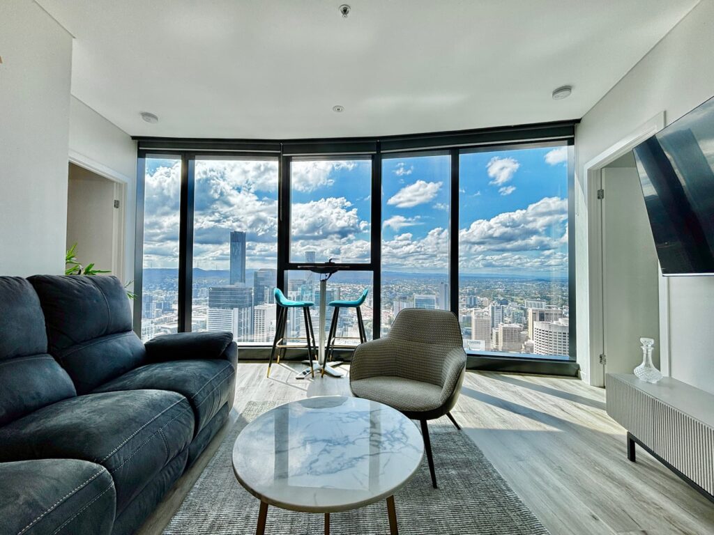 A modern living room inside the Brisbane Skytower featuring a grey sofa, marble table, and floor-to-ceiling windows with a panoramic view of the Brisbane CBD skyline and river.